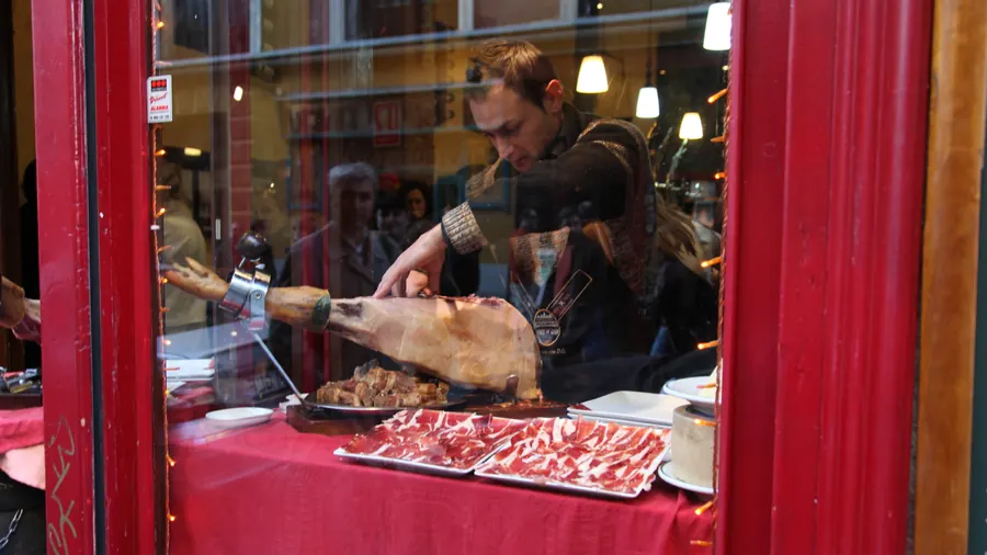 Un hombre corta jamón ibérico en un jamonero, visto a través del escaparate de un bar